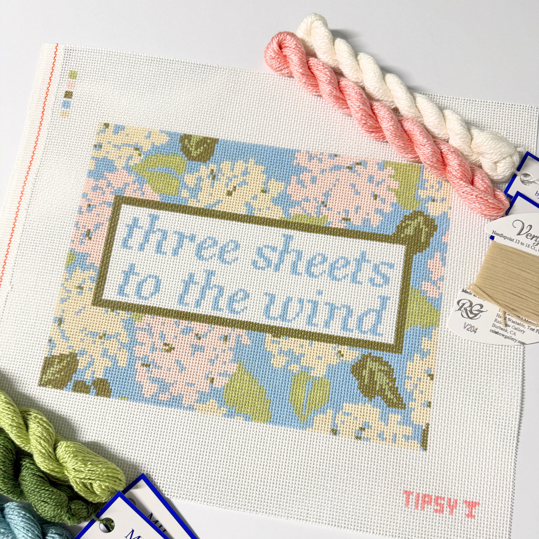 Needlepoint Kit project with 'three sheets to the wind' text and floral design, surrounded by yarn and threads.