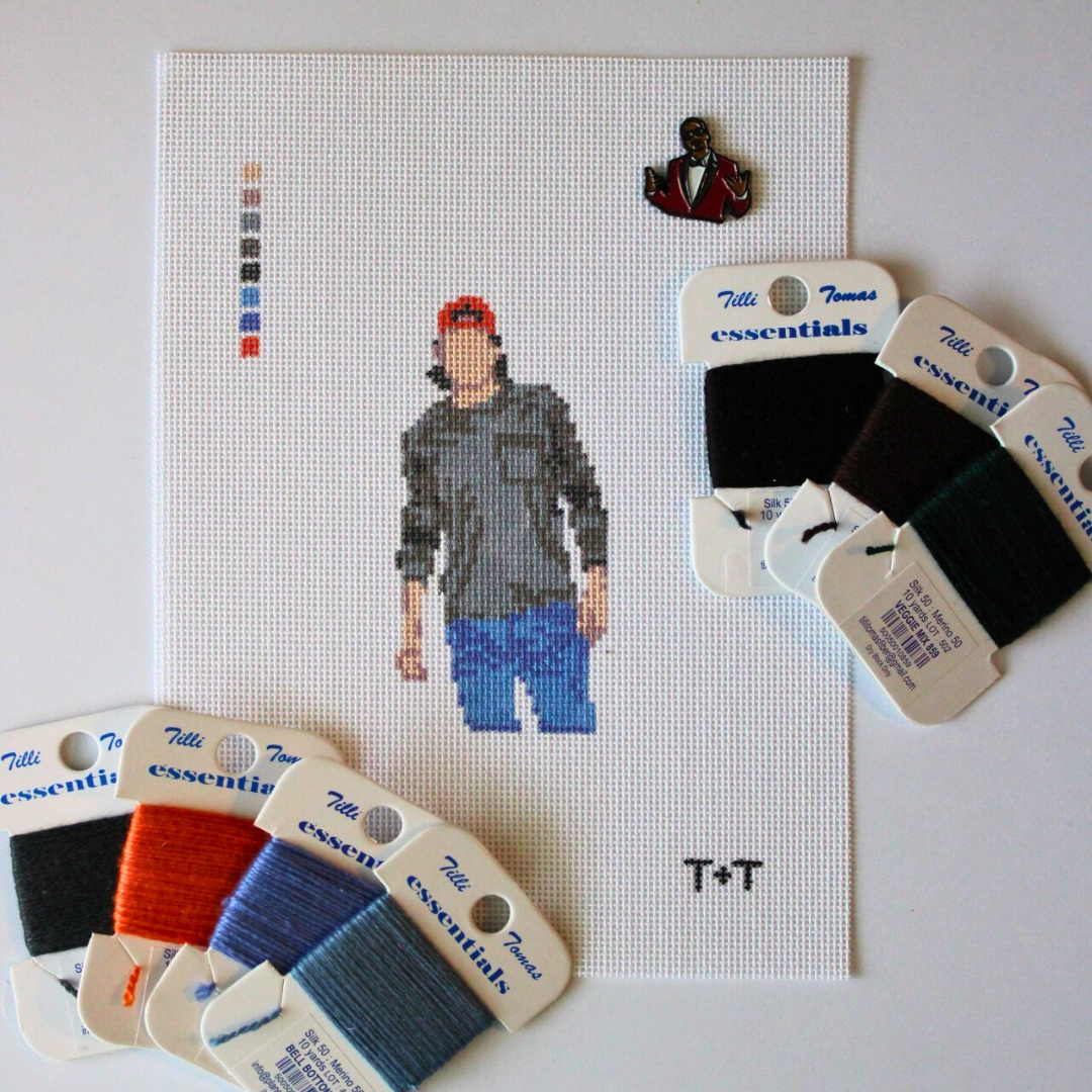 Alt text:
Threaded & Tipsy needlepoint canvas featuring a pixel-style figure of a man wearing a red baseball cap, gray shirt, and blue jeans. The canvas includes a stitched color guide and the initials "T+T" in the bottom right corner. Surrounding the canvas are six spools of Threaded & Tipsy needlepoint threads in black, brown, green, orange, and two shades of blue. A small enamel pin of a man in a red blazer is pinned near the top of the canvas.