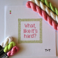 Threaded & Tipsy hand-painted needlepoint canvas featuring the phrase “what, like it’s hard?” in pink lettering, centered within a green geometric border. The initials "T+T" are painted in black in the bottom right corner. Three skeins of Threaded & Tipsy