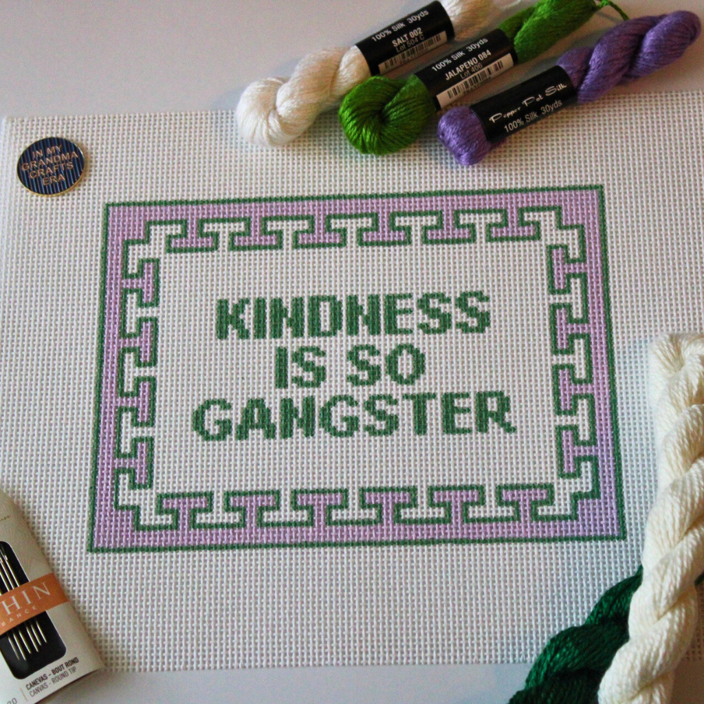 Threaded & Tipsy hand-painted needlepoint canvas featuring the phrase “KINDNESS IS SO GANGSTER” in bold green block letters, framed by a lavender and green geometric border. The canvas is surrounded by silk needlepoint threads in white, green, and purple,