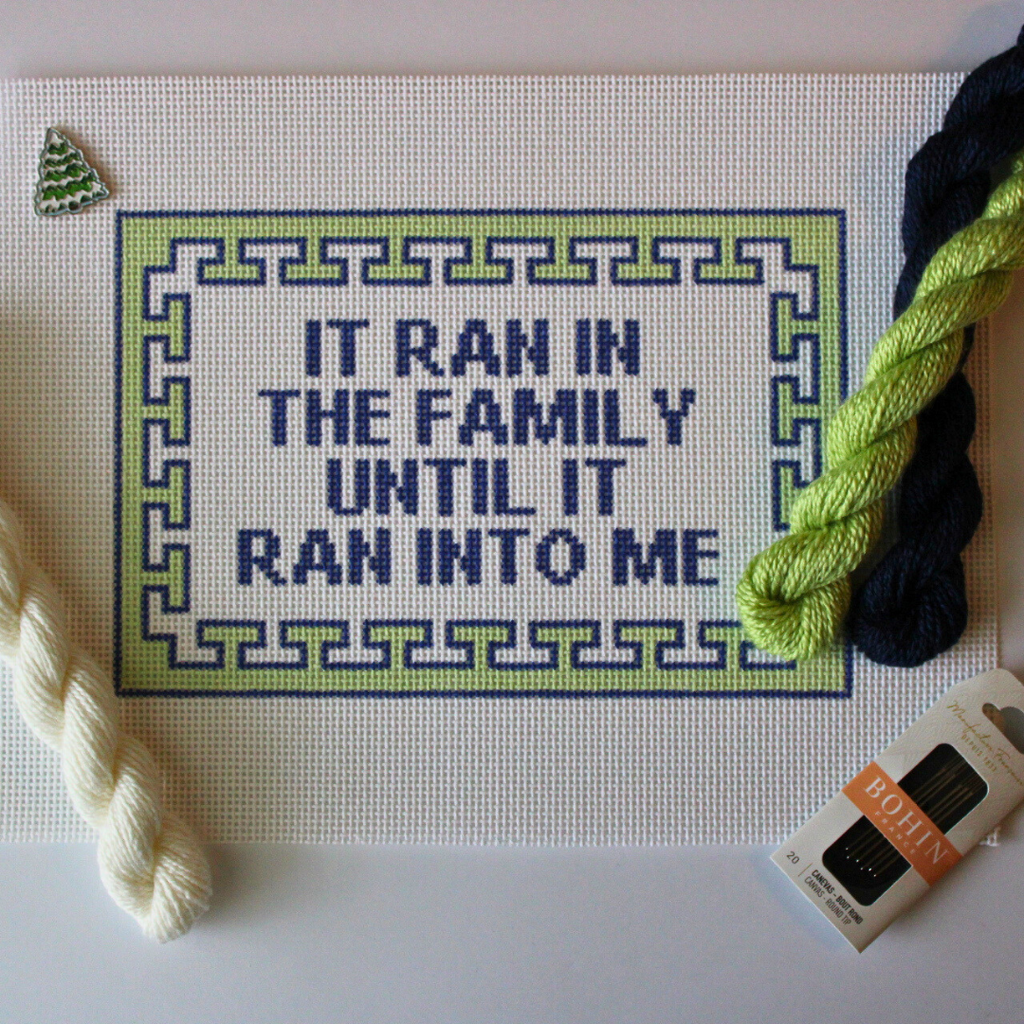 Threaded & Tipsy hand-painted needlepoint canvas featuring the phrase “IT RAN IN THE FAMILY UNTIL IT RAN INTO ME” in bold navy blue block letters. The quote is framed by a navy and lime green geometric border. Surrounding the canvas are twisted skeins of 