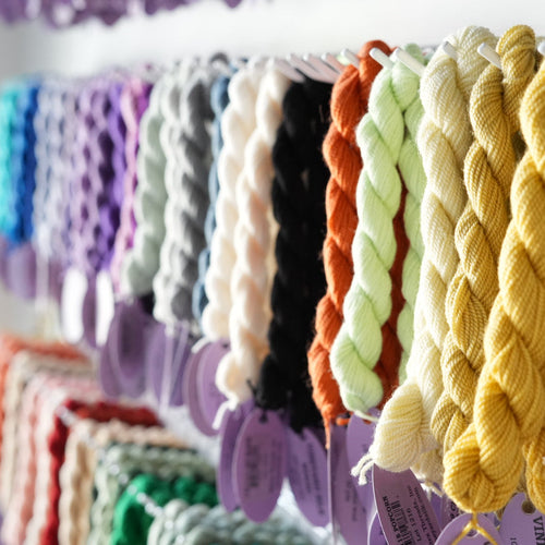 A close-up row of braided yarn skeins in multiple colors, including yellow, cream, light green, black, orange, purple, white, gray, blue, and pink, hanging on a display rack.