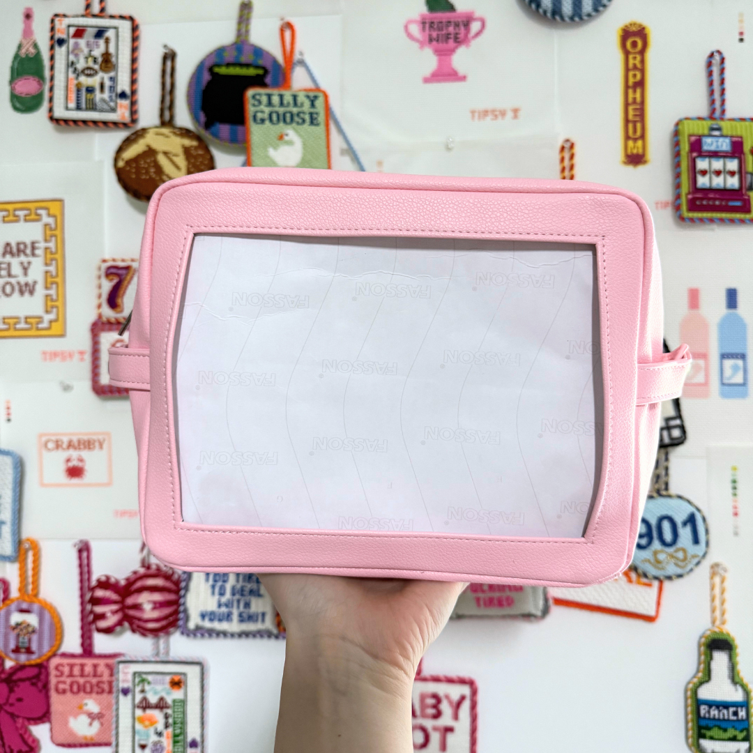 Pink self finishing needlepoint pouch in front of a colorful wall with various items.