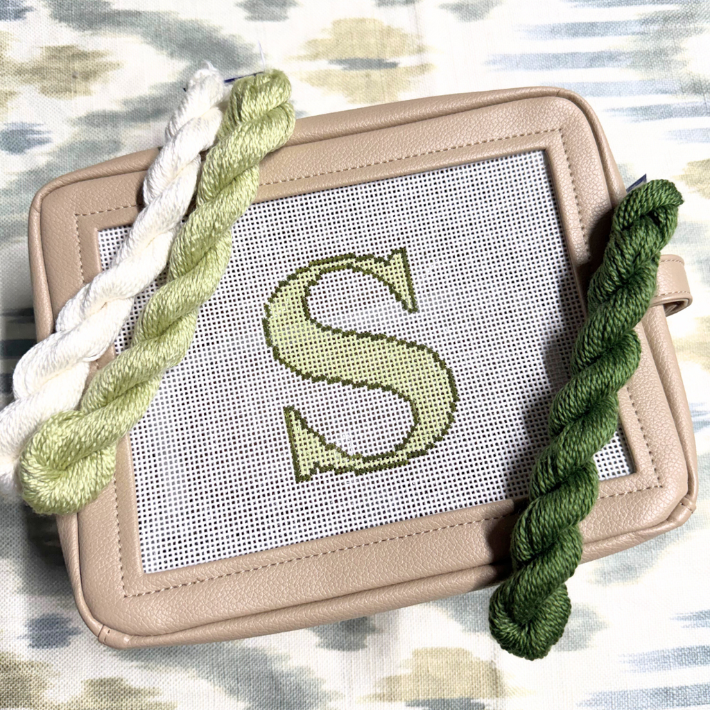 needlepoint kit with a completed letter 'S' design on canvas, surrounded by green yarn on a patterned background.